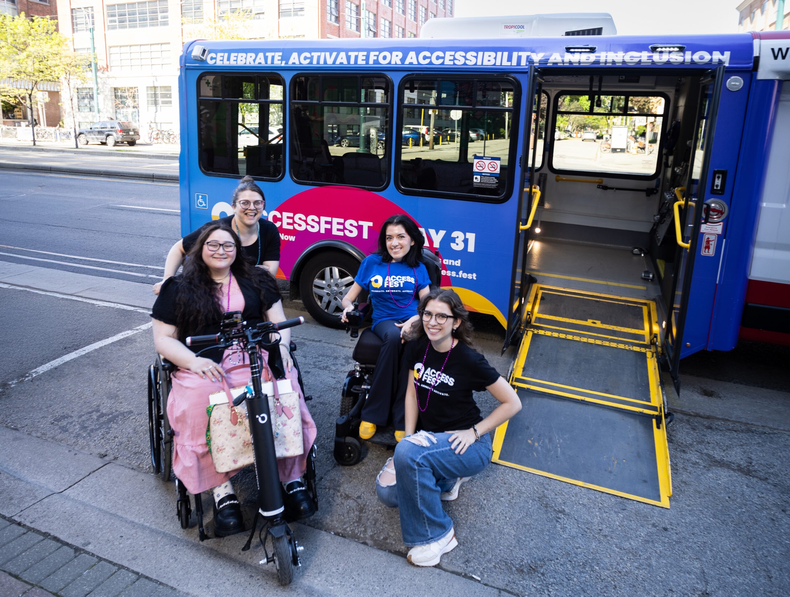 Mayaan Ziv, a passenger, and two others wearing AccessFest T-shirts pose in front of a WheelTrans bus, with text that reads "Celebrate, Activate for Accessibility and Inclusion" and "AccessFest May 31".