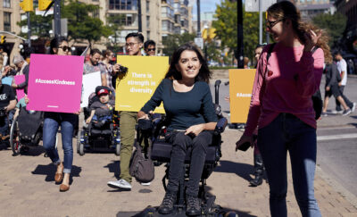 Mayaan Ziv on a sidewalk alongside other community members. In the background, people hold signs that read "#AccessKindness" and "There is strength in diversity"