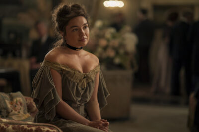 "A woman in a vintage off-shoulder dress sits on a patterned sofa in a softly-lit, elegant room with blurred people and floral arrangements in the background, suggesting a formal or historic gathering."