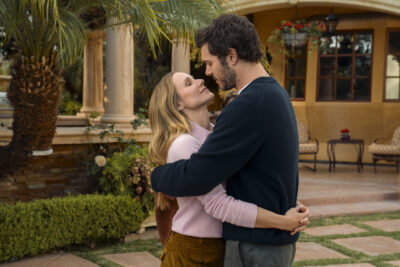 "A man and woman embrace and look into each other's eyes in a lush garden setting with palm trees, manicured bushes, and an elegant patio with chairs and columns in the background."